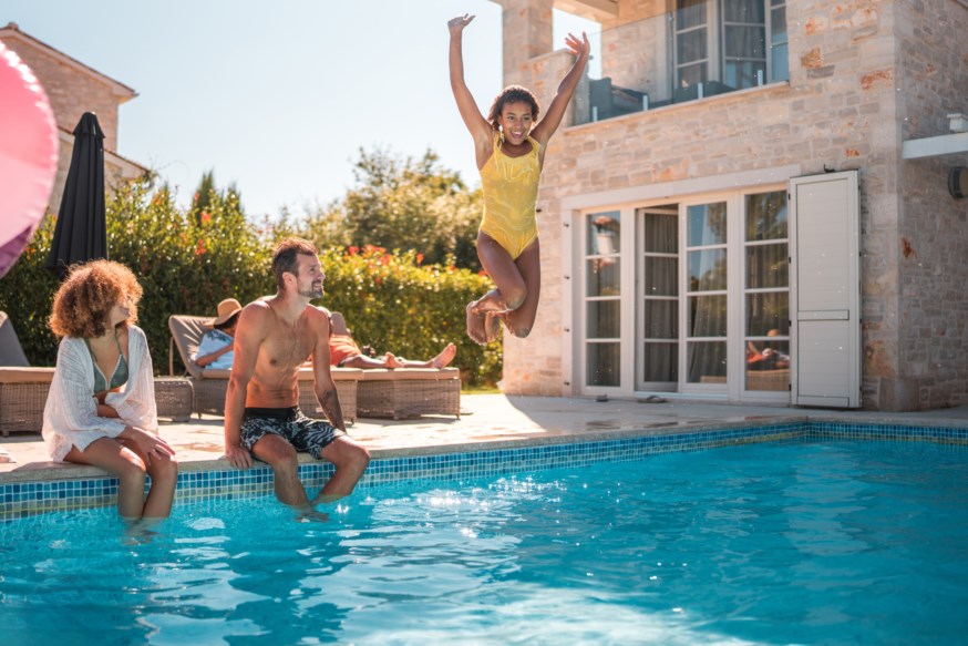 Familie og venner koser seg ved bassenget, en ung jente i gult badeantrekk hopper smilende inn i vannet.
